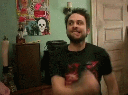 A Man With A Beard Is Standing In Front Of A Skull Poster On The Wall GIF