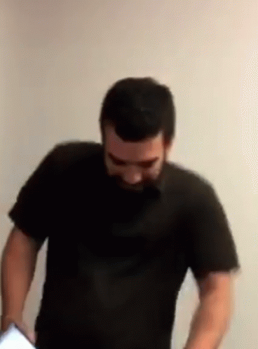 A Man With A Beard Is Standing In Front Of A White Wall Wearing A Black Shirt GIF