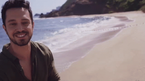 A Man With A Beard Is Standing On A Beach And Smiling GIF