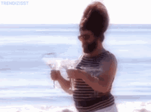 A Man With A Beard Is Standing On A Beach Holding A Martini Glass . GIF