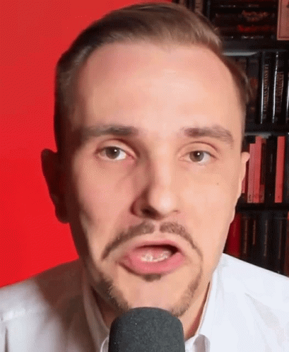 A Man With A Beard Is Talking Into A Microphone In Front Of A Bookshelf Filled With Books . GIF