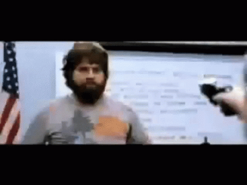 A Man With A Beard Is Talking Into A Microphone In Front Of An American Flag .. GIF