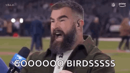 A Man With A Beard Is Talking Into A Microphone On A Sports Field . GIF