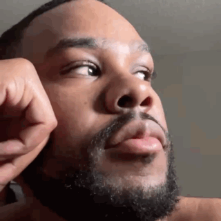 A Man With A Beard Is Touching His Face With His Hand And Looking Up . GIF