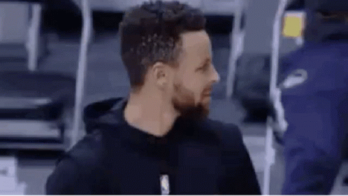 A Man With A Beard Is Wearing A Black Hoodie And Standing In Front Of A Basketball Court . GIF