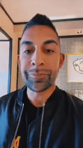 A Man With A Beard Is Wearing A Black Jacket With The Letter M On The Front GIF