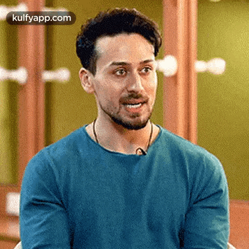 A Man With A Beard Is Wearing A Blue Shirt And Smiling While Sitting In Front Of A Mirror . GIF