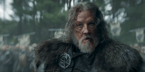 A Man With A Beard Is Wearing A Fur Coat And A Badge With A Cross On It . GIF