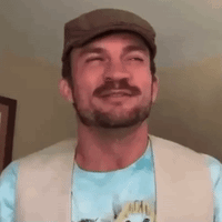 A Man With A Beard Is Wearing A Hat And A Vest And Smiling . GIF