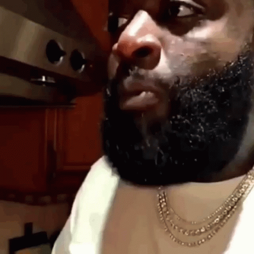 A Man With A Beard Is Wearing A Necklace And A White Shirt In A Kitchen . GIF