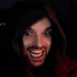 A Man With A Beard Is Wearing A Red Hoodie And Smiling In A Dark Room . GIF