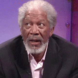 A Man With A Beard Is Wearing A Suit And A Pink Shirt And Making A Surprised Face . GIF