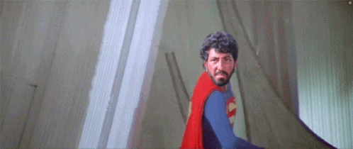 A Man With A Beard Is Wearing A Superman Costume And Standing In Front Of A Wall . GIF