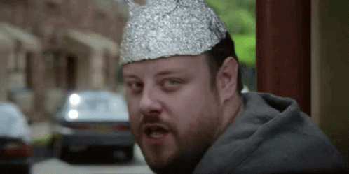 A Man With A Beard Is Wearing A Tin Foil Hat And Making A Funny Face . GIF