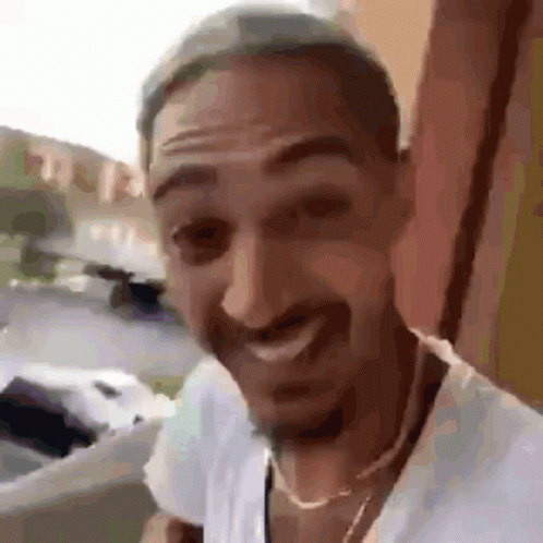 A Man With A Beard Is Wearing A White Shirt And A Gold Chain Around His Neck . GIF