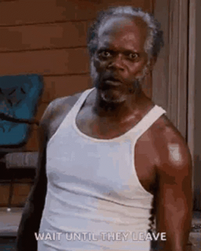 A Man With A Beard Is Wearing A White Tank Top And Standing In Front Of A Chair . GIF
