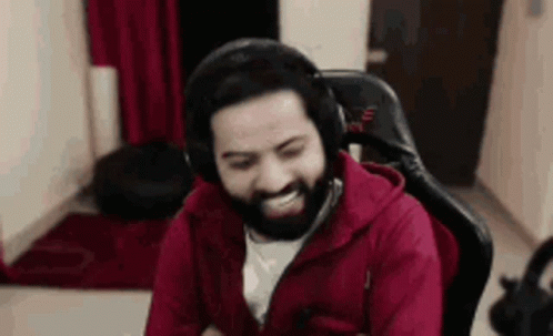 A Man With A Beard Is Wearing Headphones And A Red Jacket While Sitting In A Chair . GIF
