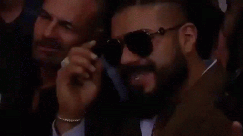 A Man With A Beard Is Wearing Sunglasses While Sitting In A Crowd Of People . GIF