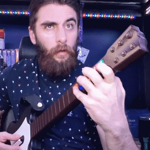 A Man With A Beard Playing A Guitar In Front Of A Bookshelf GIF