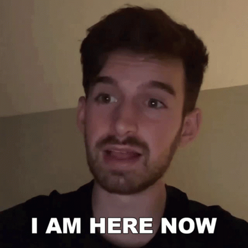 A Man With A Beard Says " I Am Here Now " In Front Of His Face GIF