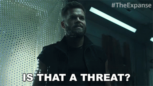 A Man With A Beard Says " Is That A Threat " In A Dark Room GIF