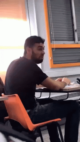 A Man With A Beard Sits At A Desk In A Classroom GIF