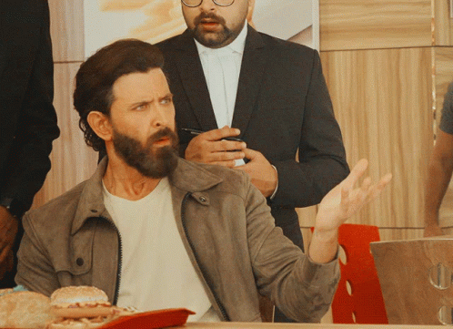 A Man With A Beard Sits At A Table With A Tray Of Hamburgers GIF
