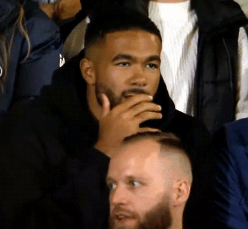 A Man With A Beard Sits In A Crowd With His Hand To His Mouth GIF