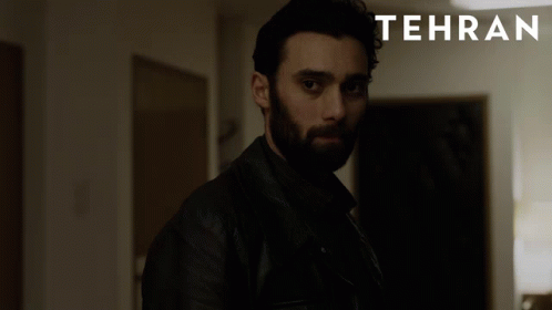 A Man With A Beard Stands In A Dark Room With Tehran Written On The Bottom GIF
