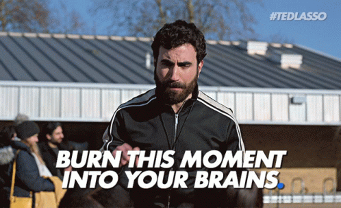 A Man With A Beard Stands In Front Of A Building With Burn This Moment Into Your Brains Written Below Him GIF