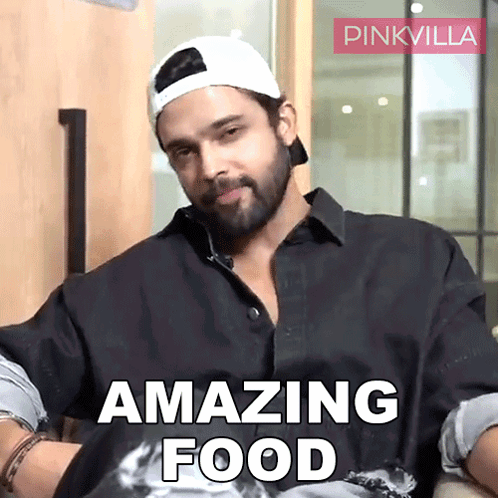 A Man With A Beard Wearing A Baseball Cap And A Black Shirt Says " Amazing Food " GIF