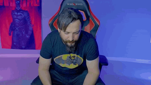A Man With A Beard Wearing A Batman Shirt Is Sitting In A Chair GIF