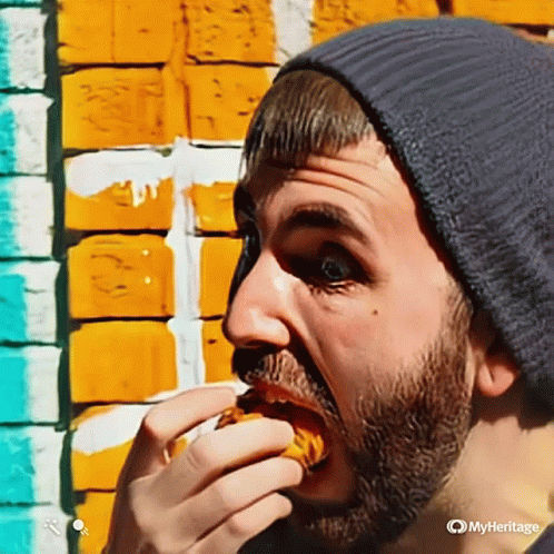 A Man With A Beard Wearing A Beanie Is Eating A Sandwich GIF
