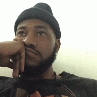 A Man With A Beard Wearing A Black Beanie Is Sitting Down With His Hand On His Face . GIF