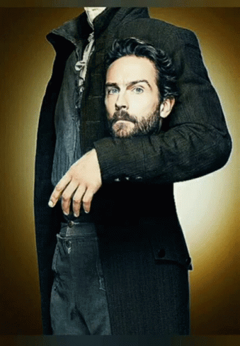 A Man With A Beard Wearing A Black Coat Has His Hand On His Pocket GIF