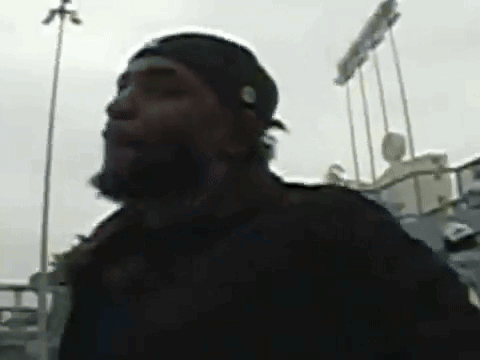 A Man With A Beard Wearing A Black Hat Is Standing In Front Of A Baseball Stadium . GIF
