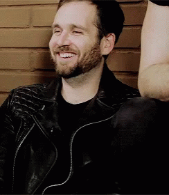A Man With A Beard Wearing A Black Leather Jacket Is Smiling While Sitting In Front Of A Brick Wall . GIF