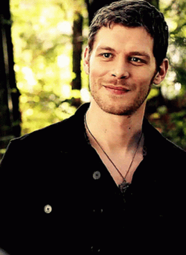 A Man With A Beard Wearing A Black Shirt And A Necklace Smiles For The Camera GIF