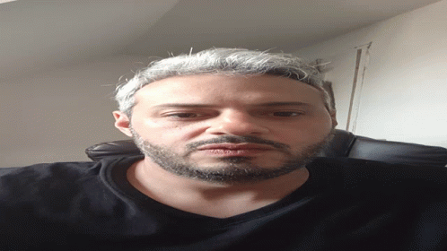 A Man With A Beard Wearing A Black Shirt Looks Down At The Camera GIF