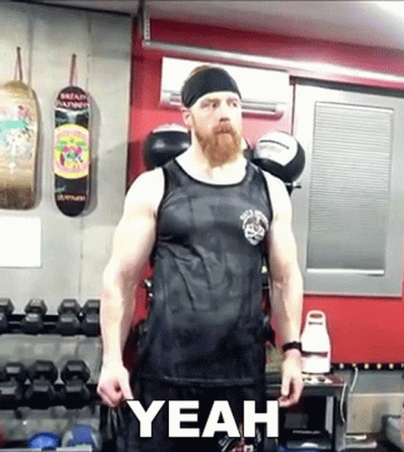 A Man With A Beard Wearing A Black Tank Top Is Standing In A Gym With The Word Yeah Written On The Bottom GIF
