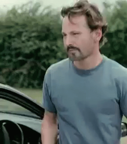 A Man With A Beard Wearing A Blue Shirt Is Standing Next To A Car . GIF
