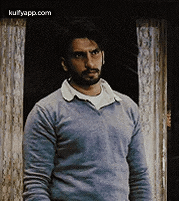A Man With A Beard Wearing A Blue Sweater Is Standing In Front Of A Door . GIF