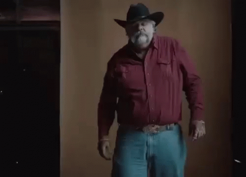 A Man With A Beard Wearing A Cowboy Hat And Gloves Is Standing In Front Of A Brown Wall . GIF