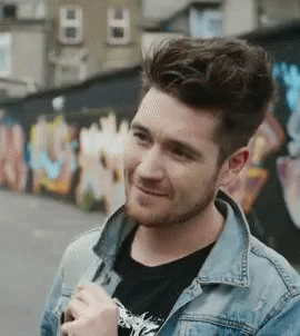 A Man With A Beard Wearing A Denim Jacket And A Black Shirt Is Smiling In Front Of A Graffiti Wall . GIF