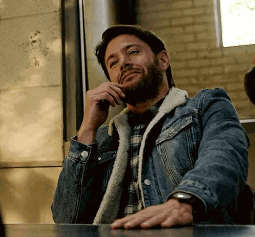 A Man With A Beard Wearing A Denim Jacket Is Sitting At A Table Talking On A Cell Phone . GIF