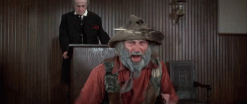 A Man With A Beard Wearing A Feathered Hat Stands In Front Of A Judge GIF