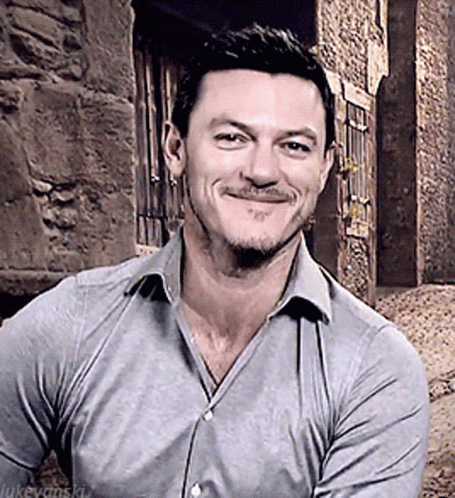 A Man With A Beard Wearing A Grey Shirt Is Smiling In Front Of A Stone Wall GIF