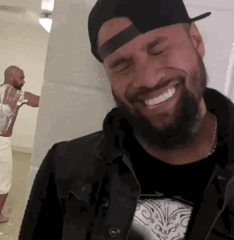 A Man With A Beard Wearing A Hat And A Black Jacket Is Smiling While Another Man Is Standing Behind Him . GIF