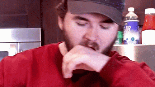 A Man With A Beard Wearing A Hat And A Red Shirt Eating GIF