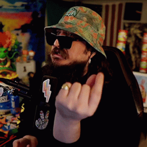 A Man With A Beard Wearing A Hat And Sunglasses Is Sitting In Front Of A Microphone Giving The Middle Finger . GIF